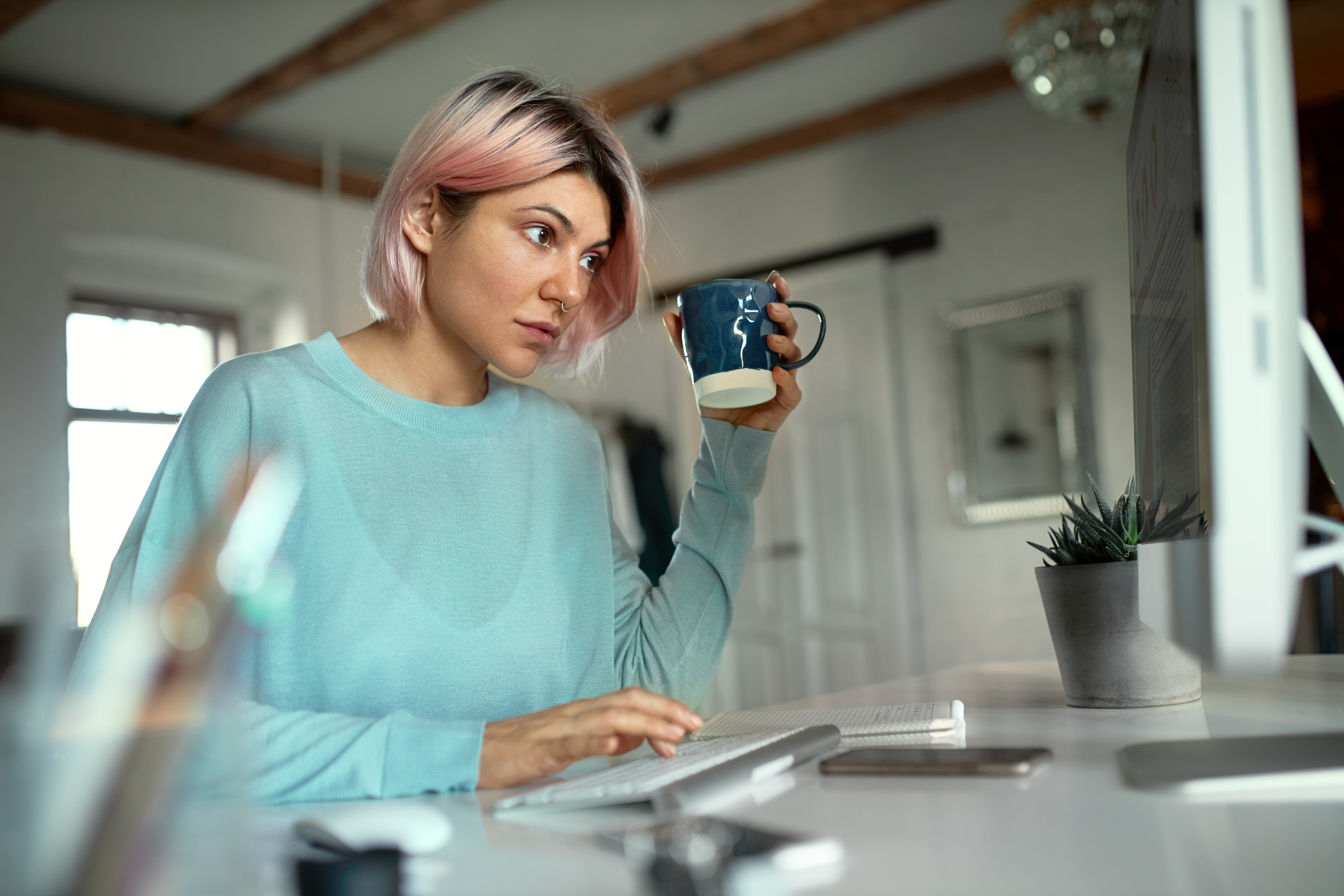 Young female with pink hair sitting at her desk typing and drinking tea